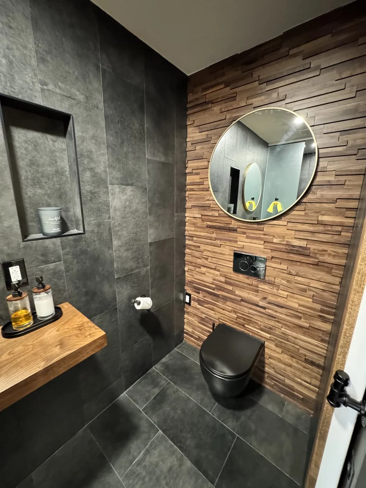 Modern bathroom remodel with a round mirror, wood accent wall, and dark tile